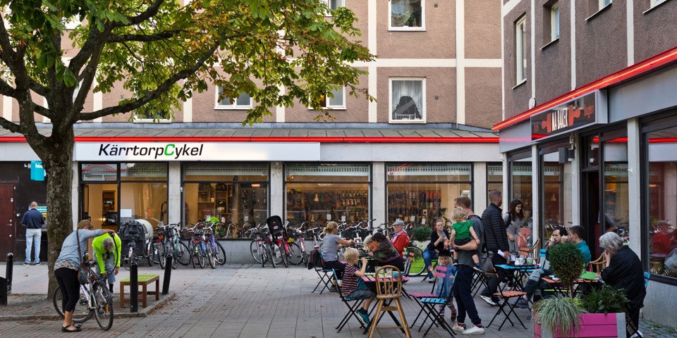 Ett litet torg med ett café som har uteservering och ligger bredvid en cykelverkstad.