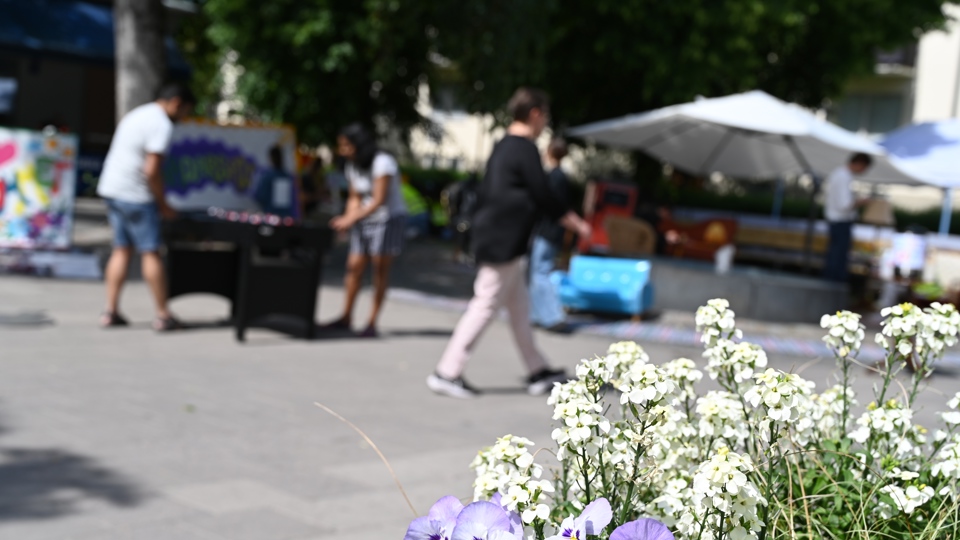 Människor som promenerar över torget och två människor som spelar ett bordsfotbollsspel på torget. På torget finns det planteringar, sittplatser under parasoll och träd. Foto.