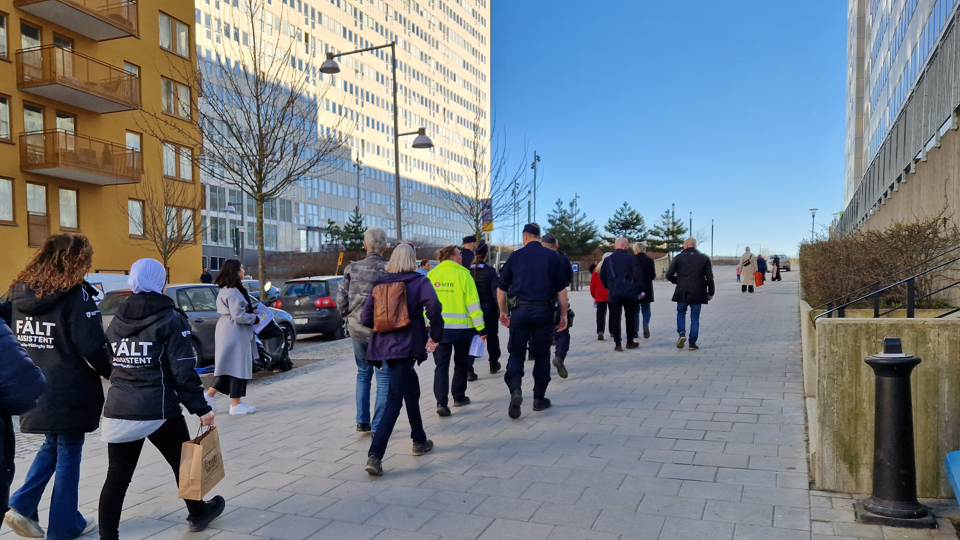 Flera personer genomför en trygghetsvandring bland höga flerfamiljshus, även fältassistent och polis medverkar.