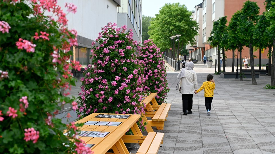 En vuxen och ett barn promenerar hand i hand på etttorg med blommor och gula bänkar.