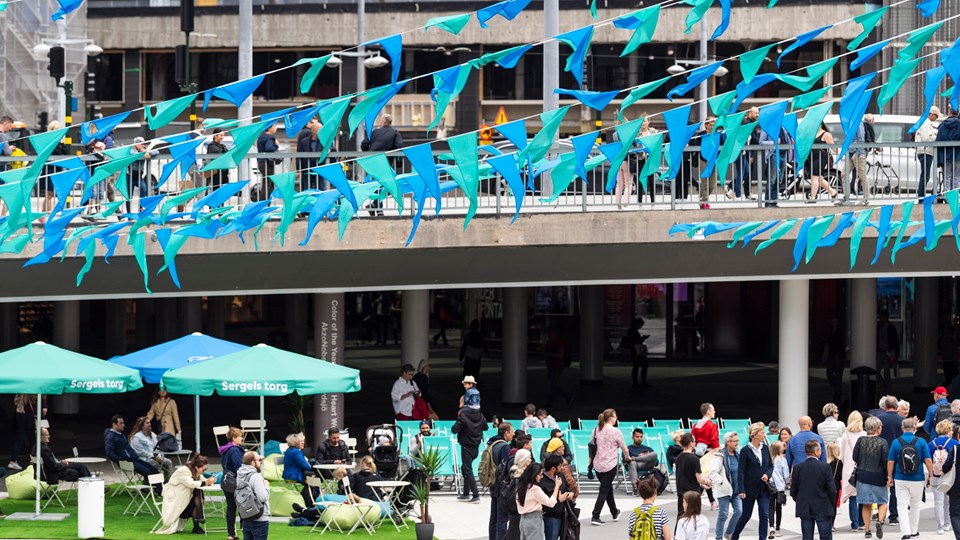 Det pågår firande på Sergels torg. Färgglada blå och gröna vimplar hänger över torget. Det vimlar av människor på torget och samtal pågår.