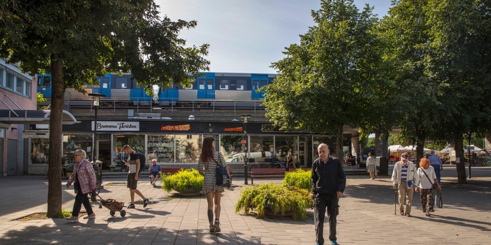 Torg på Brommaplan. Människor som promenerar över torget. Tunnelbana som kör på tunnelbanespår som löper ovanför torget. På torget finns det planterade träd, planteringar, affärer och fik. Foto.