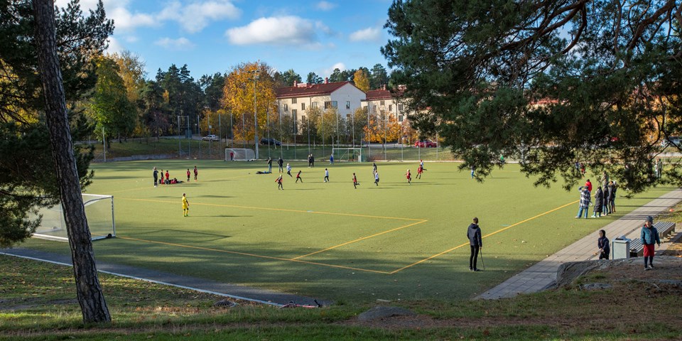 Fotbollsmatch mellan två lag på en fotbollsplan. Runt fotbollsplanen finns det skog och omkringliggande hus. Foto.