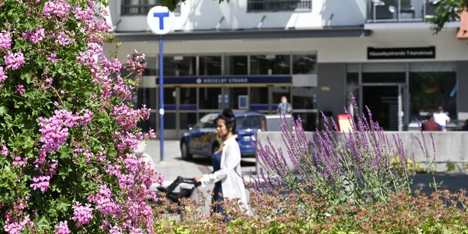 Blommor och en kvinna som drar en barnvagn i solen vid en tunnelbanenedgång.