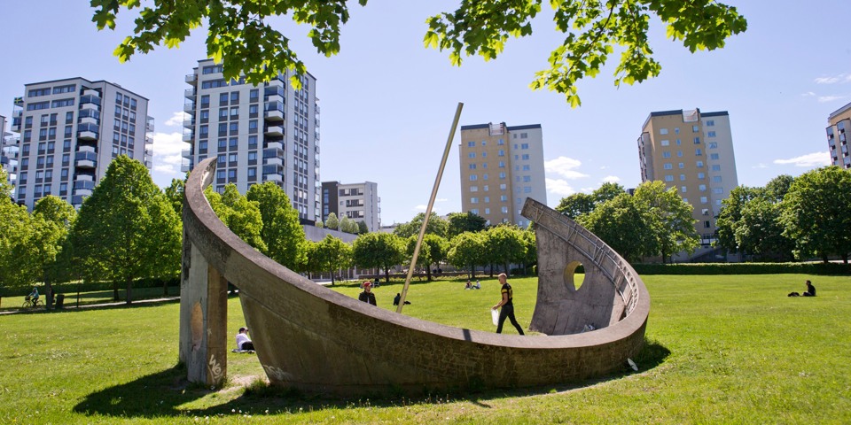 En stor halvcirkelformad betonginstallation i en stor park med höga flerfamiljshus i bakgrunden.