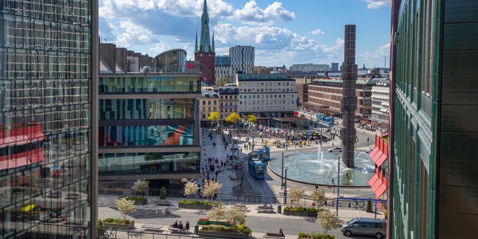 Vy mellan husen över Sergels torg och fontänen. Gatorna runt Sergels torg trafikeras av bilar och spårvagn.  Torget myllrar av människor.