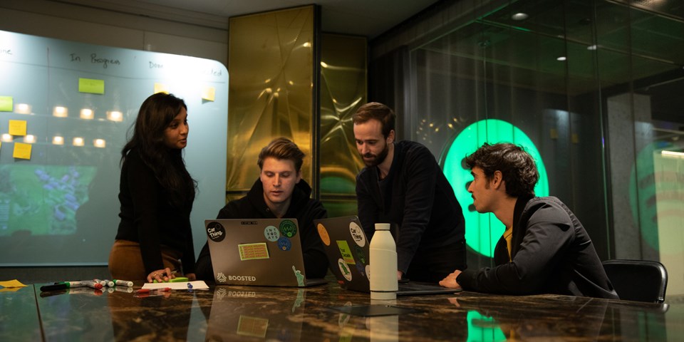 Fyra personer tittar på en datorskärm. I bakgrunden syns en digital whiteboard med post-it-lappar. DAtorn står på ett spegelblankt bord i en modernt rum.