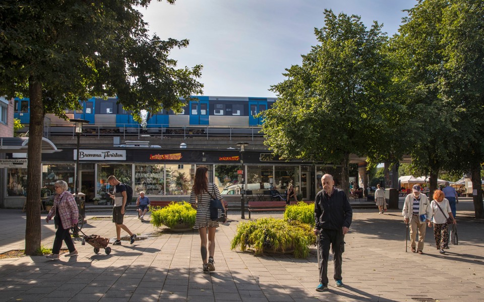 Torg på Brommaplan. Människor som promenerar över torget. Tunnelbana som kör på tunnelbanespår som löper ovanför torget. På torget finns det planterade träd, planteringar, affärer och fik. Foto.