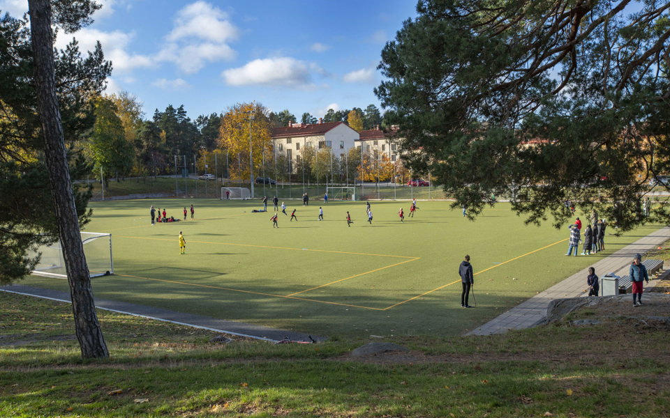Fotbollsmatch mellan två lag på en fotbollsplan. Runt fotbollsplanen finns det skog och omkringliggande hus. Foto.