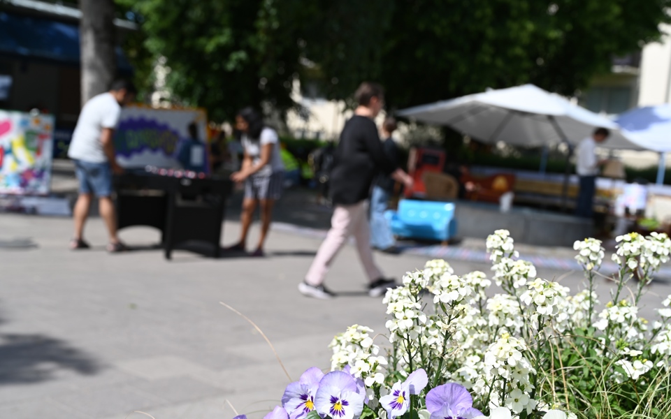Människor som promenerar över torget och två människor som spelar ett bordsfotbollsspel på torget. På torget finns det planteringar, sittplatser under parasoll och träd. Foto.