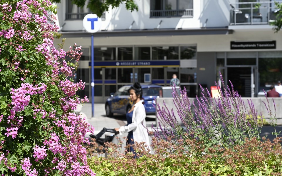 Blommor och en kvinna som drar en barnvagn i solen vid en tunnelbanenedgång.