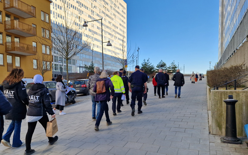 Flera personer genomför en trygghetsvandring bland höga flerfamiljshus, även fältassistent och polis medverkar.