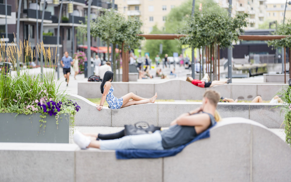 Torg med planteringar och murar där människor ligger och vilar eller läser. Människor som promenerar och springer. Foto.