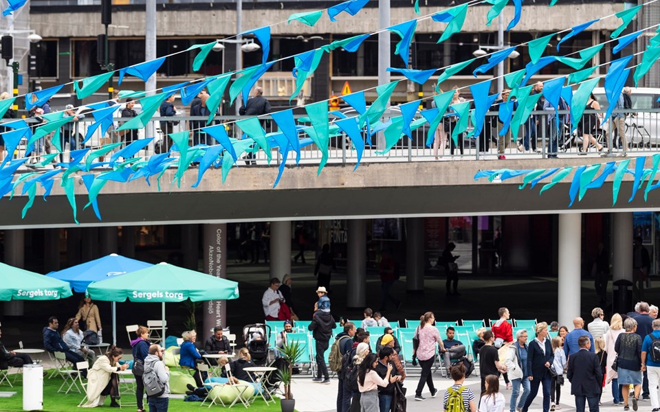 Det pågår firande på Sergels torg. Färgglada blå och gröna vimplar hänger över torget. Det vimlar av människor på torget och samtal pågår.