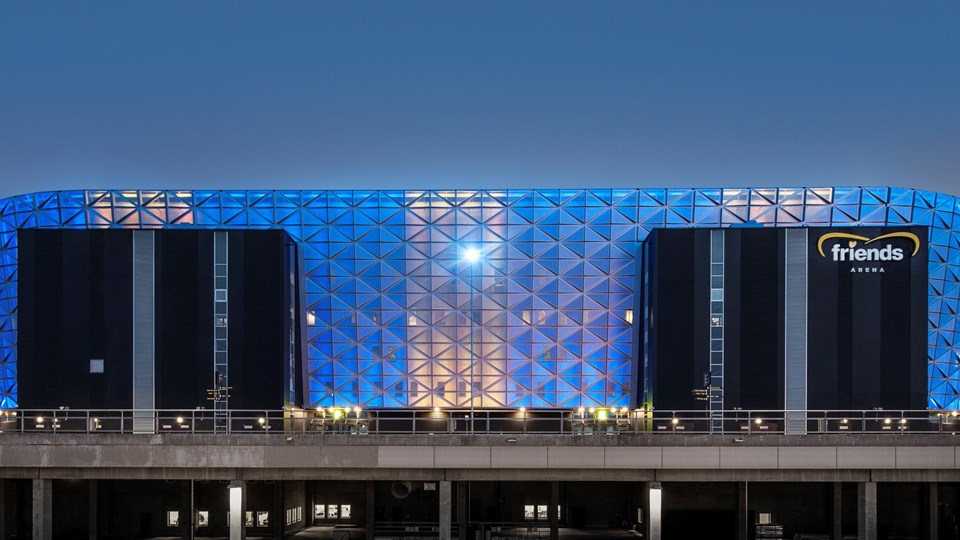 En stor arena sedd från sidan. Byggnaden lyser inifrån i blått och gult. Till högerpå arenan står texten "friends arena".
